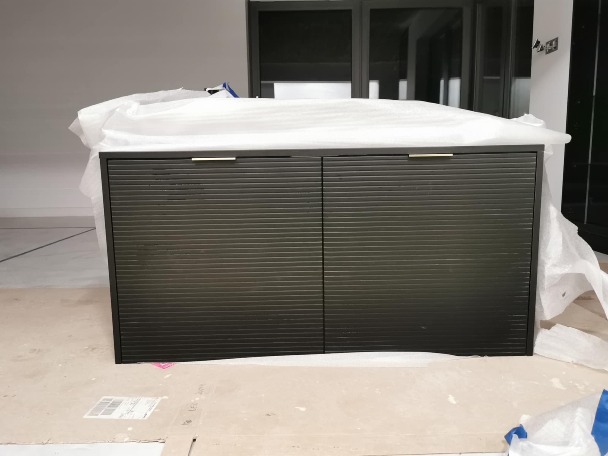 Ciemna szafka z lamelowymi drzwiami, częściowo owinięta folią ochronną, stoi na kartonie w jasnym pomieszczeniu z dużymi oknami.