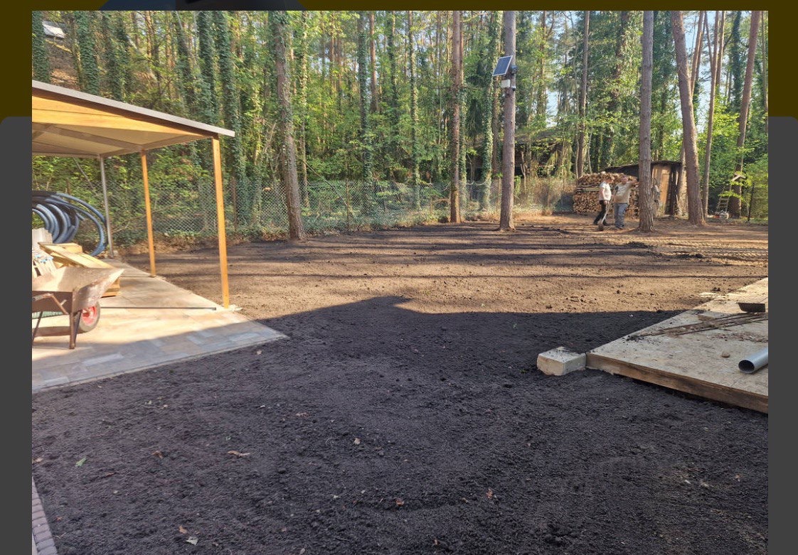 Przygotowany teren pod trawnik, świeżo wyrównana ziemia, widoczne drzewa w tle. Część terenu utwardzona z wózkiem i narzędziami. Prace ziemne w toku.