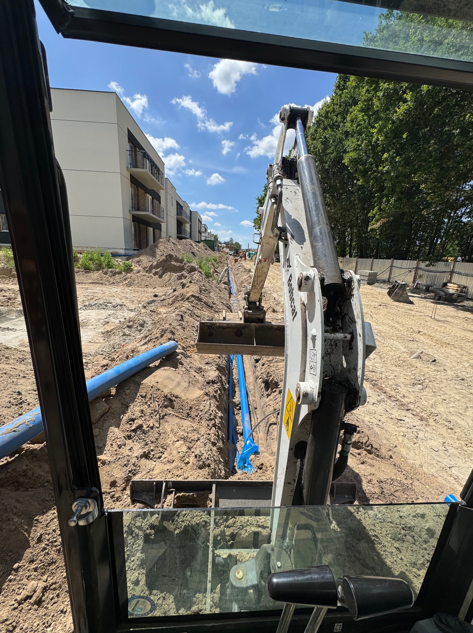 Wykop pod instalacje z widokiem z kabiny Bobcata. W tle budynki mieszkalne i błękitne rury. Wykop wąski i głęboki, przygotowany pod ułożenie instalacji.