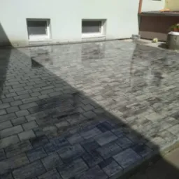 Ułożona, szara kostka brukowa przed budynkiem z dwoma oknami. Na kostce odbija się światło, a w tle widoczny element studni z kwiatami.