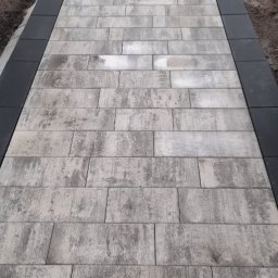 Zakład Usług BRUK-JAN TROCHOWSKI JAN - Ułożona ścieżka z kostki brukowej w odcieniach szarości, obramowana czarnym obrzeżem. W tle widoczne przygotowania do dalszych prac ziemnych.