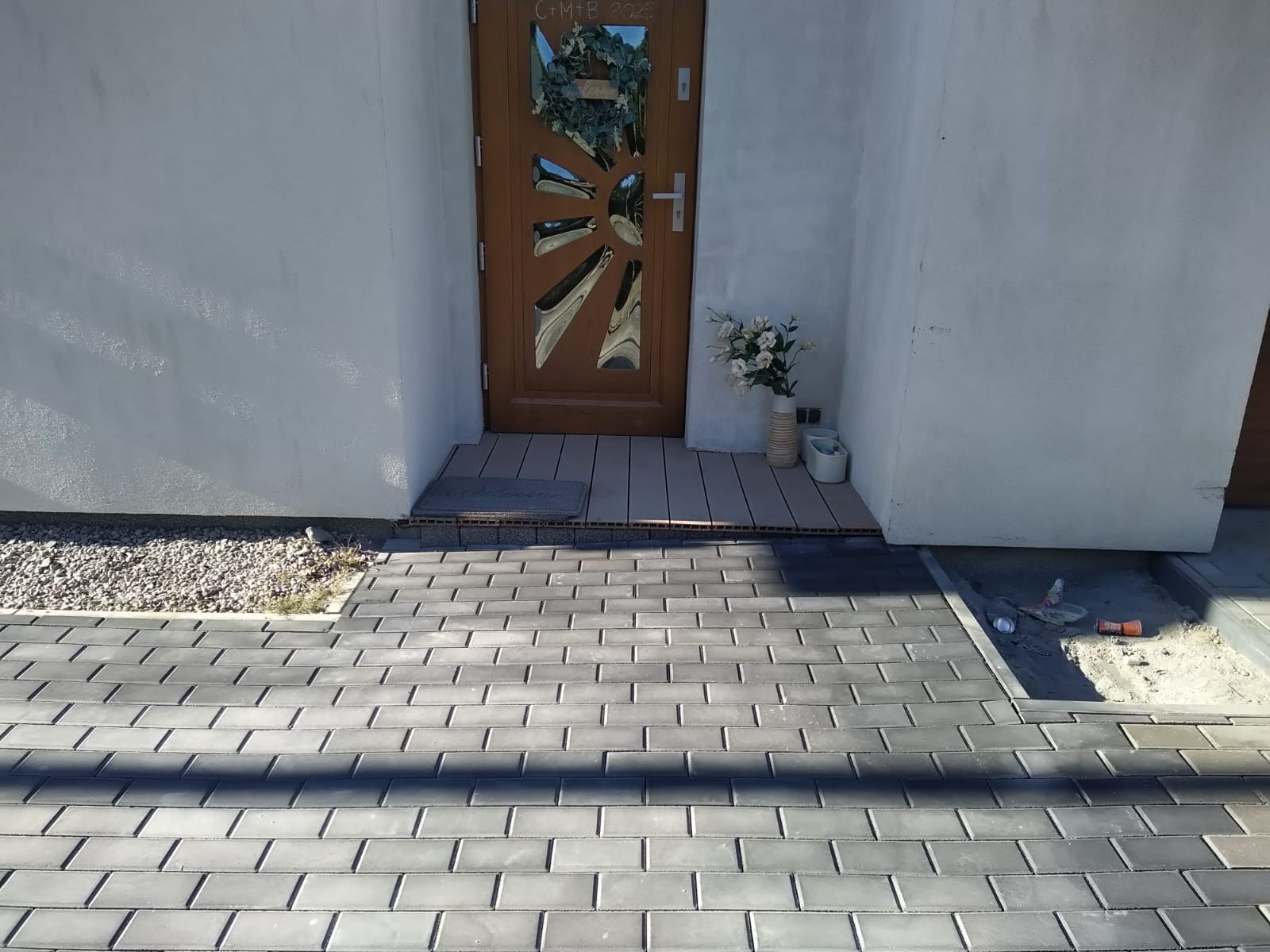 Ułożona szara kostka brukowa prowadzi do drzwi domu z dekoracjami. Widoczny fragment rabaty z kamykami i narzędzia do układania kostki.