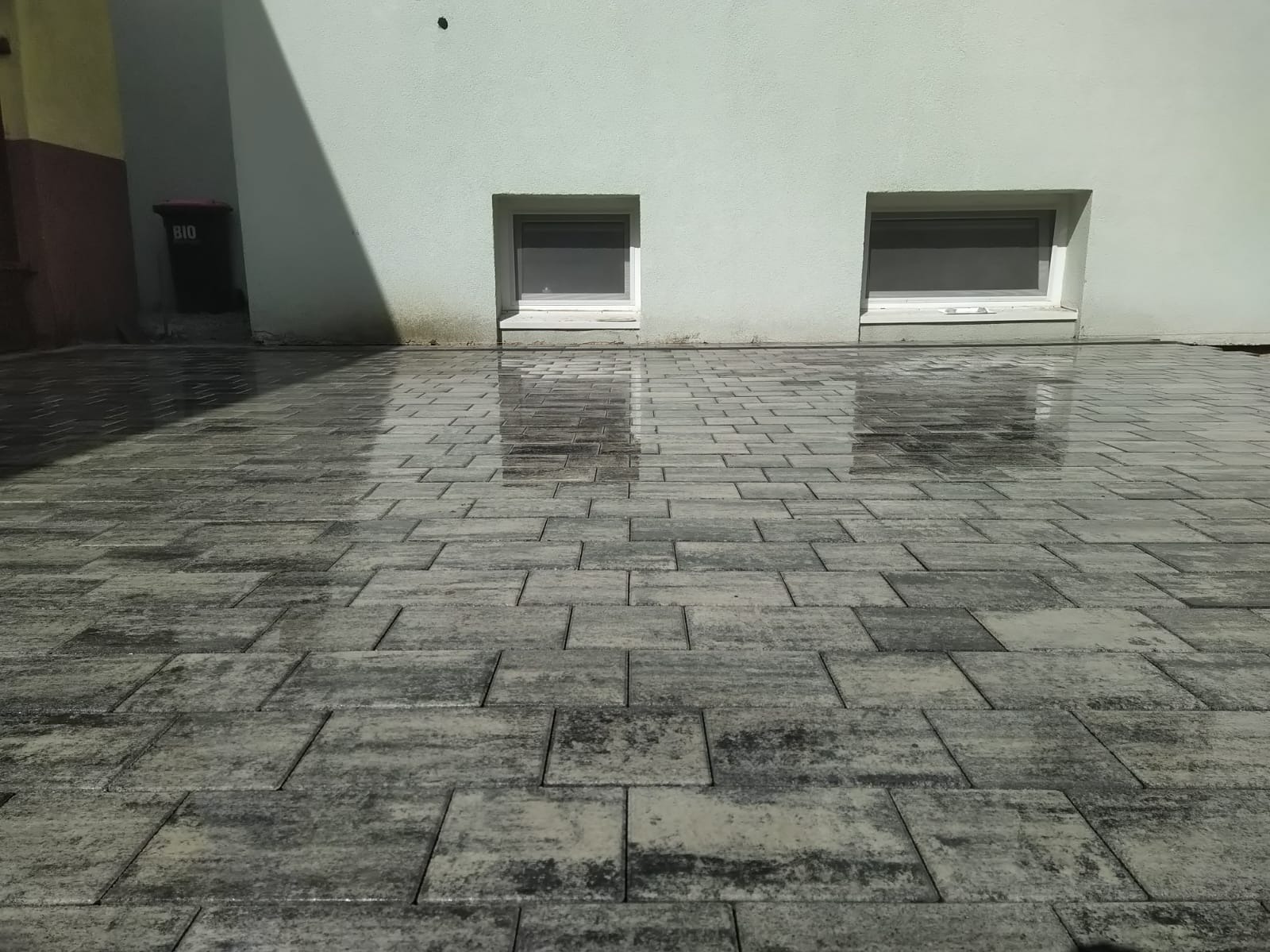 Świeżo ułożona, mokra kostka brukowa w odcieniach szarości przed budynkiem z dwoma małymi oknami. Widoczny cień i kosz na śmieci.