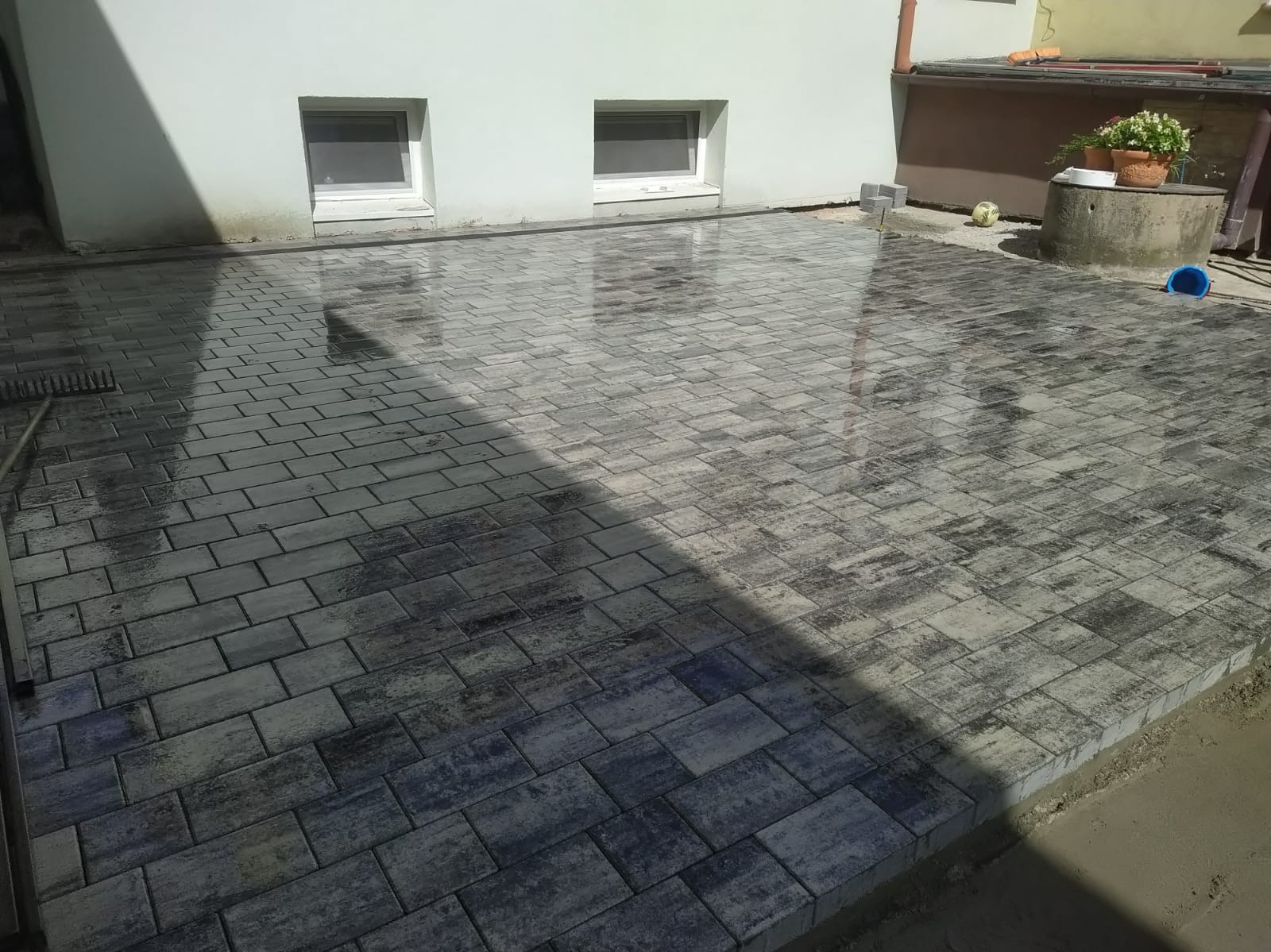 Ułożona, szara kostka brukowa przed budynkiem z dwoma oknami. Na kostce odbija się światło, a w tle widoczny element studni z kwiatami.