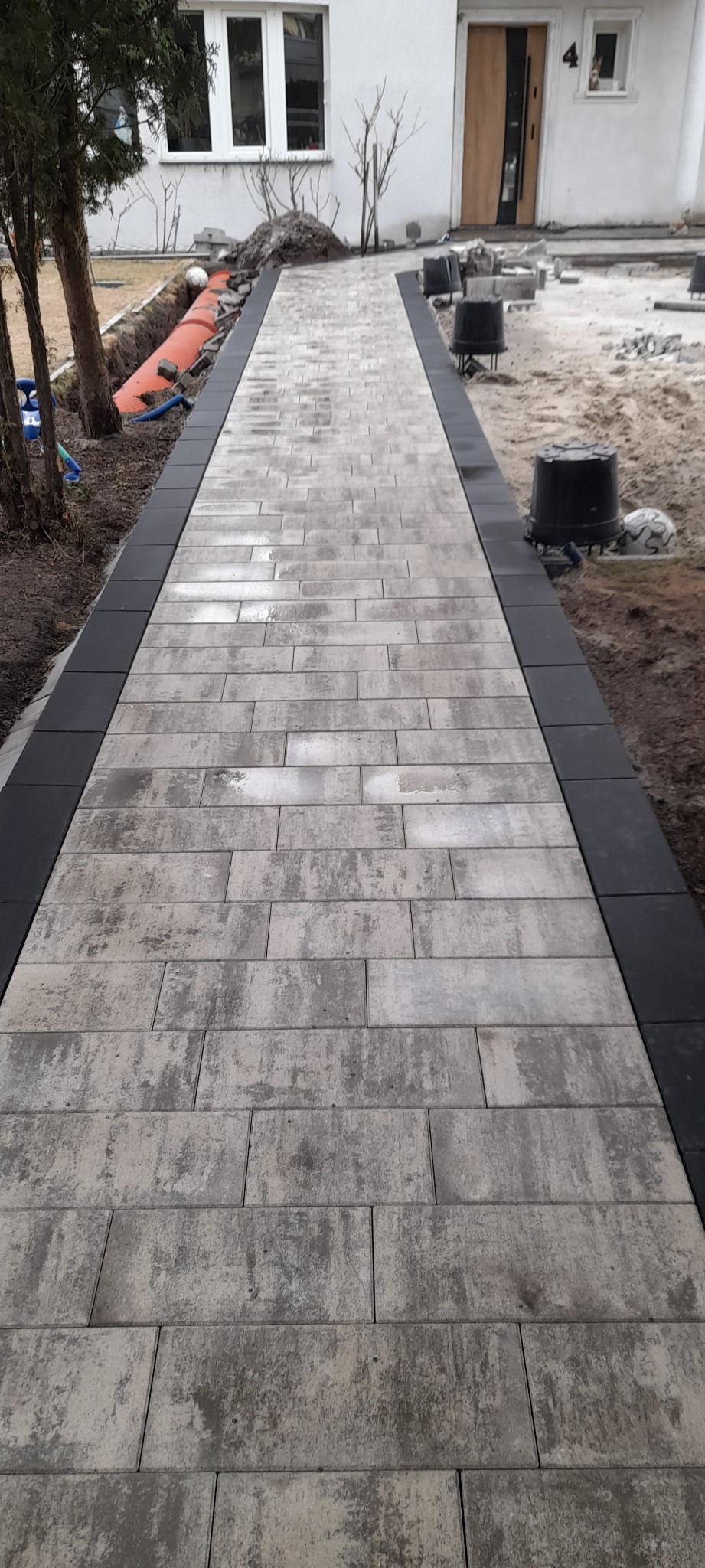 Ułożony chodnik z kostki brukowej w odcieniach szarości, obrzeża z ciemnej kostki. W tle dom z numerem 4 i fragment ogrodu z widocznymi pracami ziemnymi.