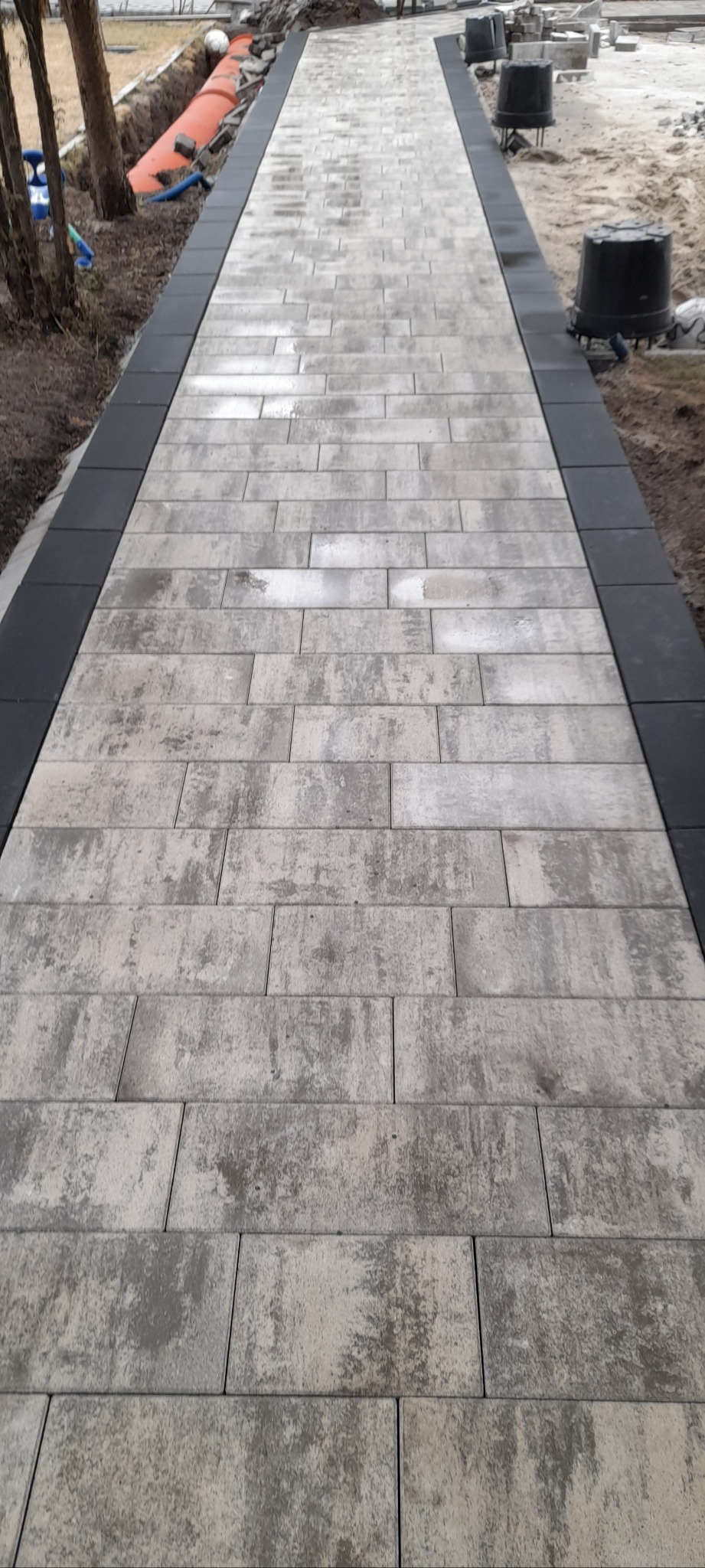 Ułożona ścieżka z kostki brukowej w odcieniach szarości, obramowana czarnym obrzeżem. W tle widoczne przygotowania do dalszych prac ziemnych.