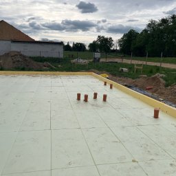 Rewelacyjny Dom - Płyta fundamentowa z jasnymi panelami izolacyjnymi XPS i wystającymi pomarańczowymi rurami instalacyjnymi, otoczona żółtą izolacją, na tle budowanego domu.