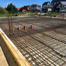 Rewelacyjny Dom - Gęsta siatka zbrojeniowa fundamentu pod budowę domu, z wystającymi pomarańczowymi rurami instalacyjnymi i drewnianymi szalunkami, w słoneczny dzień na tle osiedla.