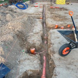 Rewelacyjny Dom - Wykopane rowy fundamentowe z ułożonymi pomarańczowymi rurami kanalizacyjnymi i niebieskim peszlem, obok stosy zbrojenia, żółtych płyt XPS i taczka.