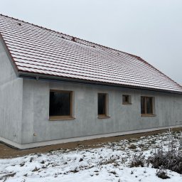 Rewelacyjny Dom - Jednopiętrowy dom z szarą elewacją i czerwonym dachem pokrytym cienką warstwą śniegu, z drewnianymi oknami, na tle zimowego krajobrazu.