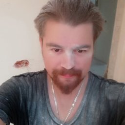 USŁUGI REMONTOWO BUDOWLANE SŁAWOMIR KWASIK - Portret mężczyzny z rudą brodą i srebrnym łańcuszkiem z krzyżykiem, pokrytego pyłem na tle ściany w trakcie prac remontowych, widoczne kable elektryczne.