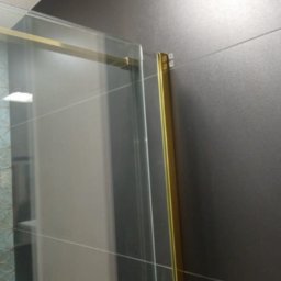 USŁUGI REMONTOWO BUDOWLANE SŁAWOMIR KWASIK - Fragment kabiny prysznicowej z złotymi profilami na tle ciemnych płytek ceramicznych z fugami.