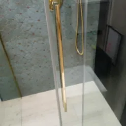 Szklana kabina prysznicowa z długim złotym uchwytem, widoczna mozaika na ścianie i złoty wąż prysznicowy.