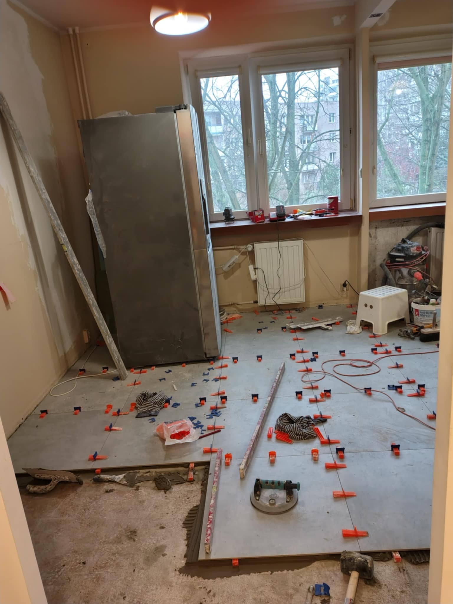 Układanie jasnoszarych płytek podłogowych w pokoju, z pomarańczowymi i niebieskimi klinami poziomującymi, widoczny przyssawka do przenoszenia płytek i młotek gumowy, w tle okno i lodówka.