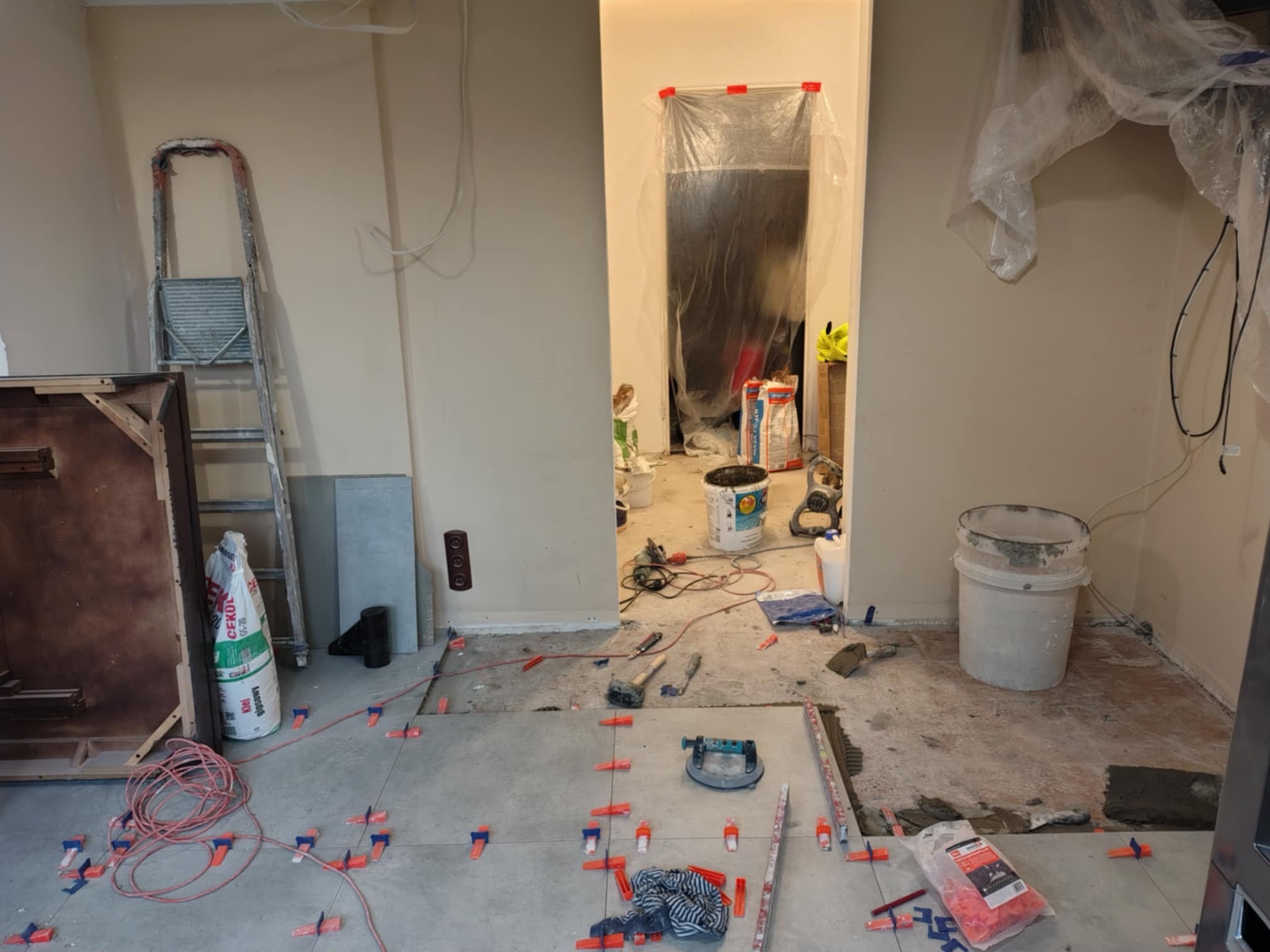 Ułożone płytki podłogowe z krzyżykami dystansowymi, widoczne narzędzia glazurnicze, drabina, worek z klejem i wiadro, pomieszczenie w trakcie remontu.