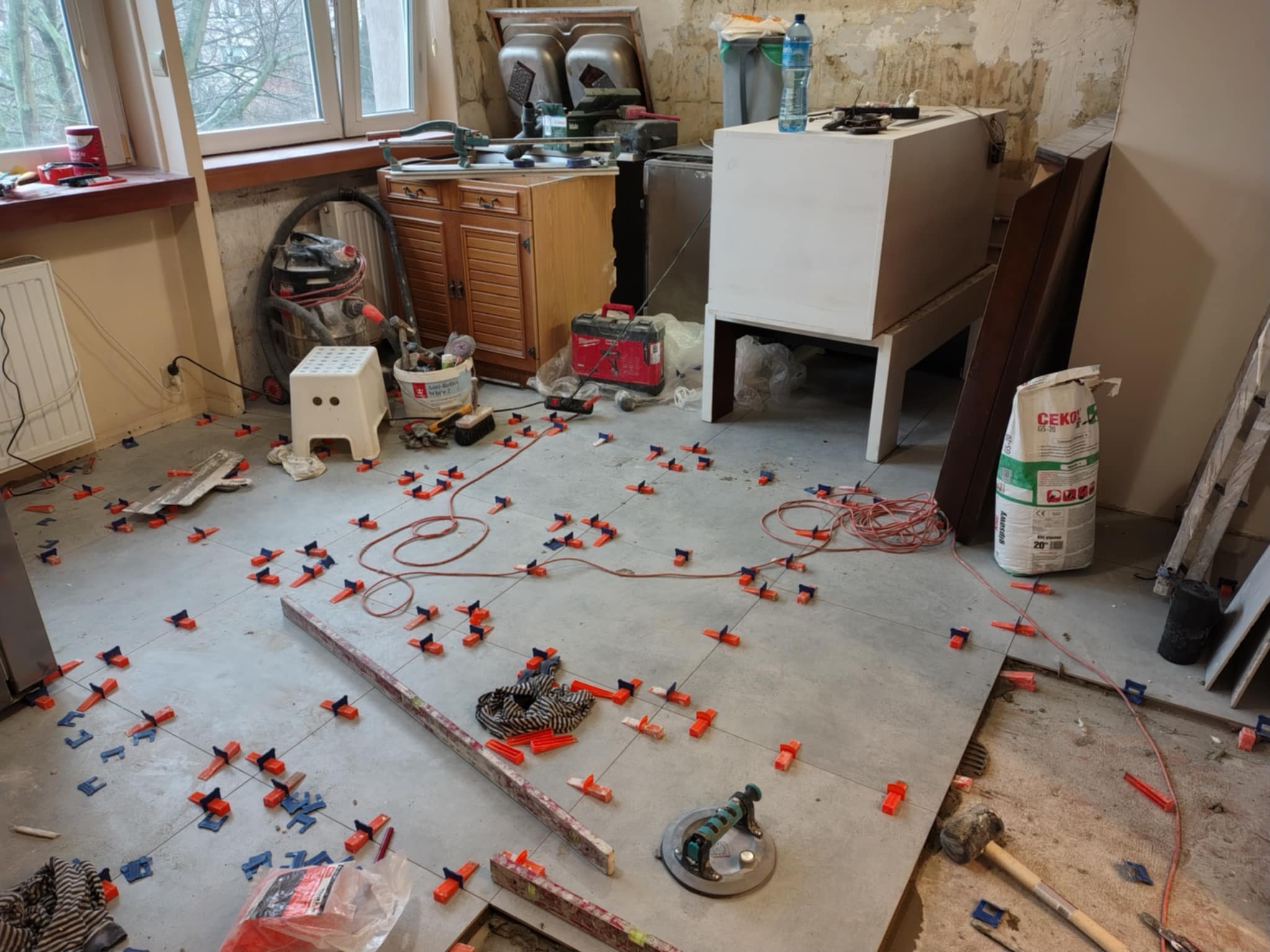 Podłoga w trakcie układania płytek, widoczne krzyżyki dystansowe, przyssawka do płytek, poziomica, młotek gumowy, worek z zaprawą klejową CEKOL, oraz nieporządek w remontowanym pomieszczeniu.