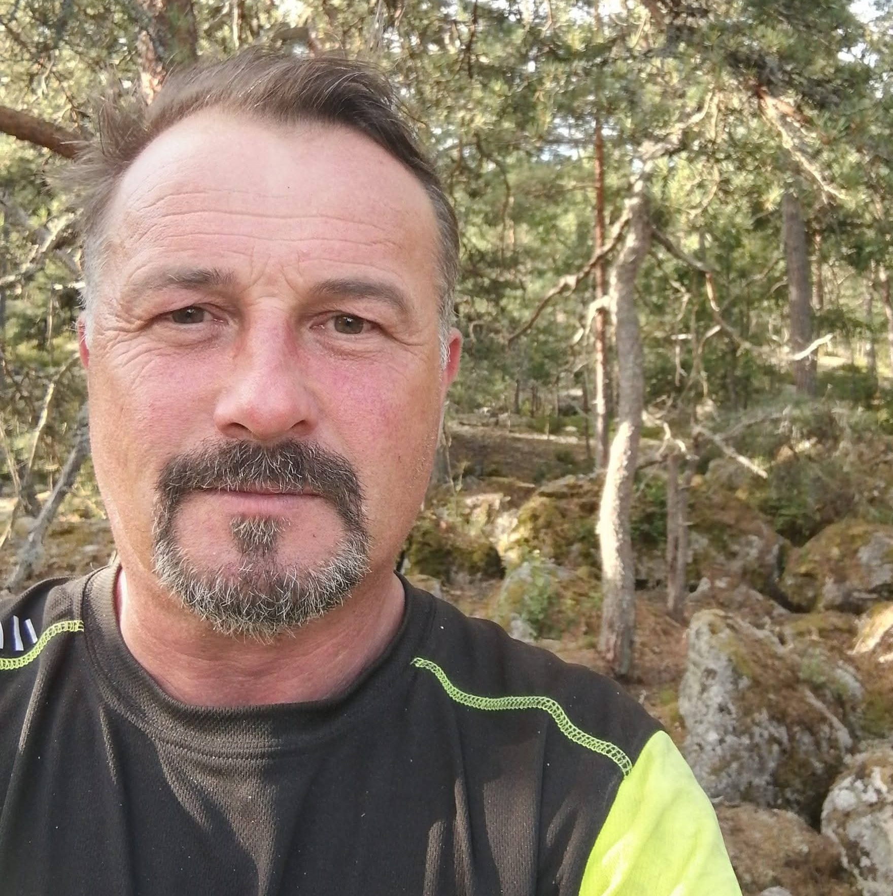 Mężczyzna z brodą i wąsami w czarno-żółtej koszulce na tle leśnych skał i drzew.