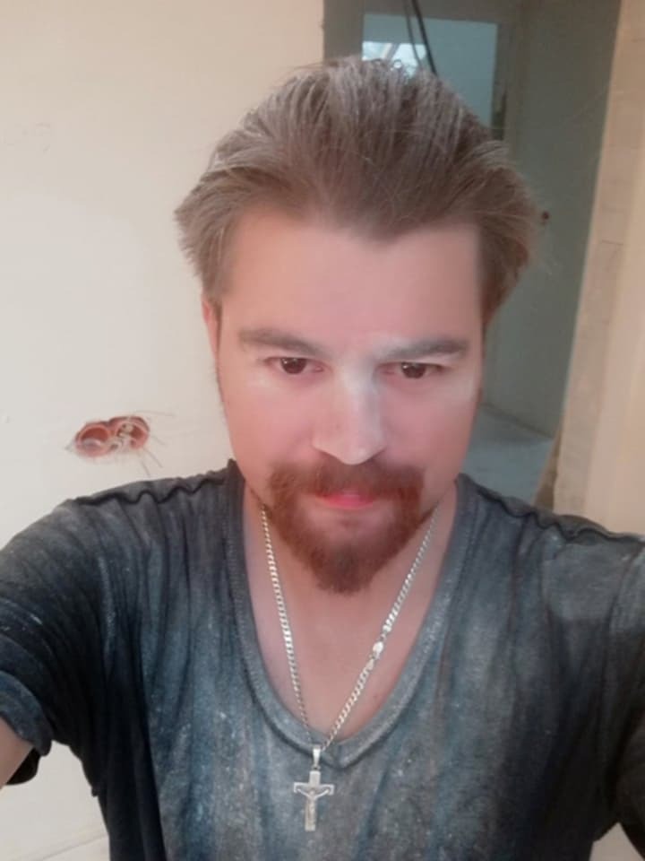 Portret mężczyzny z rudą brodą i srebrnym łańcuszkiem z krzyżykiem, pokrytego pyłem na tle ściany w trakcie prac remontowych, widoczne kable elektryczne.