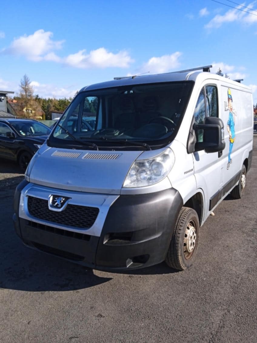 Srebrny samochód dostawczy Peugeot z grafiką przedstawiającą postać w niebieskim kombinezonie i kasku, zaparkowany na zewnątrz w słoneczny dzień.