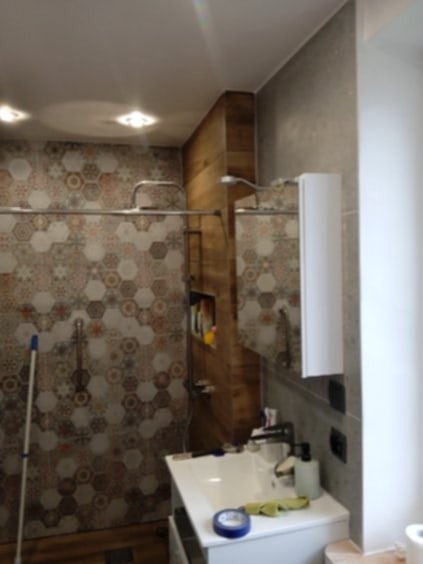 Wykończona łazienka z heksagonalnymi płytkami w odcieniach brązu i szarości, kabina prysznicowa z drewnianą wnęką na kosmetyki, umywalka z białą szafką i lustrem.