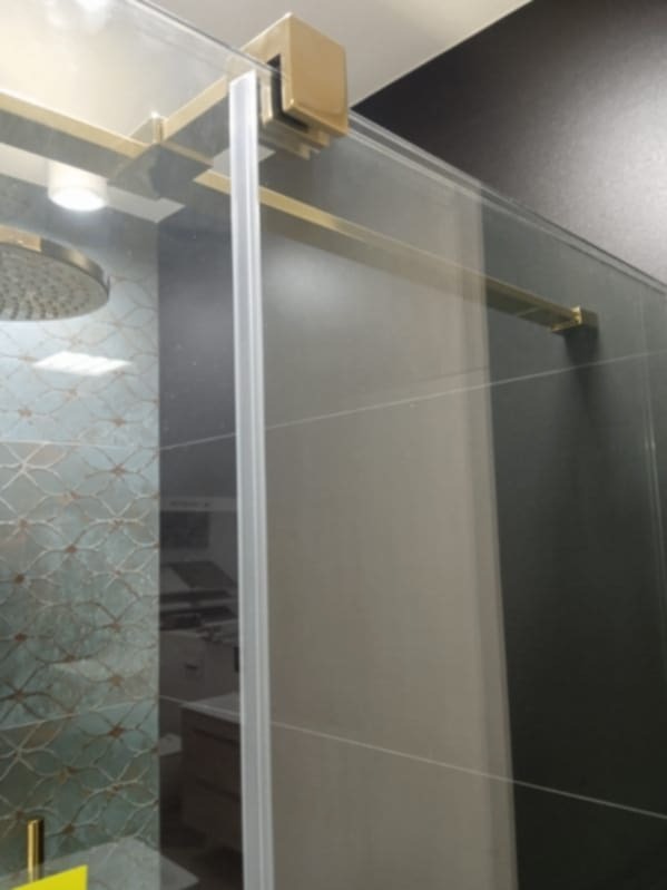 Szklana kabina prysznicowa z przesuwnymi drzwiami, złote okucia, dekoracyjne płytki na ścianie w tle.