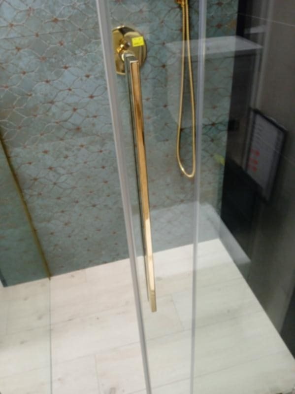 Szklana kabina prysznicowa z długim złotym uchwytem, widoczna mozaika na ścianie i złoty wąż prysznicowy.