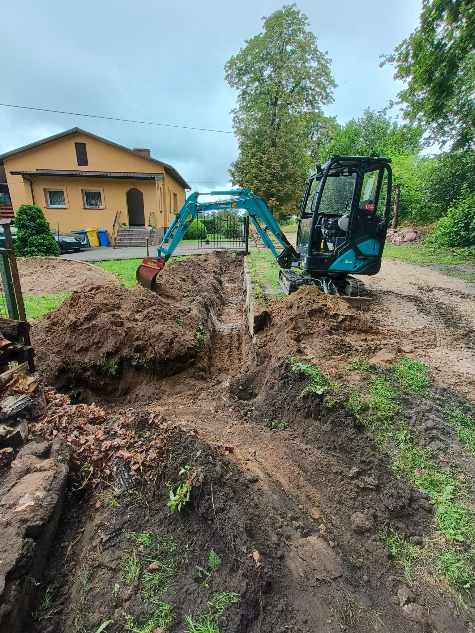 Niebieska minikoparka na posesji podczas wykopu wąskiego rowu; widoczny dom z żółtą elewacją i ogrodzenie z bramą.