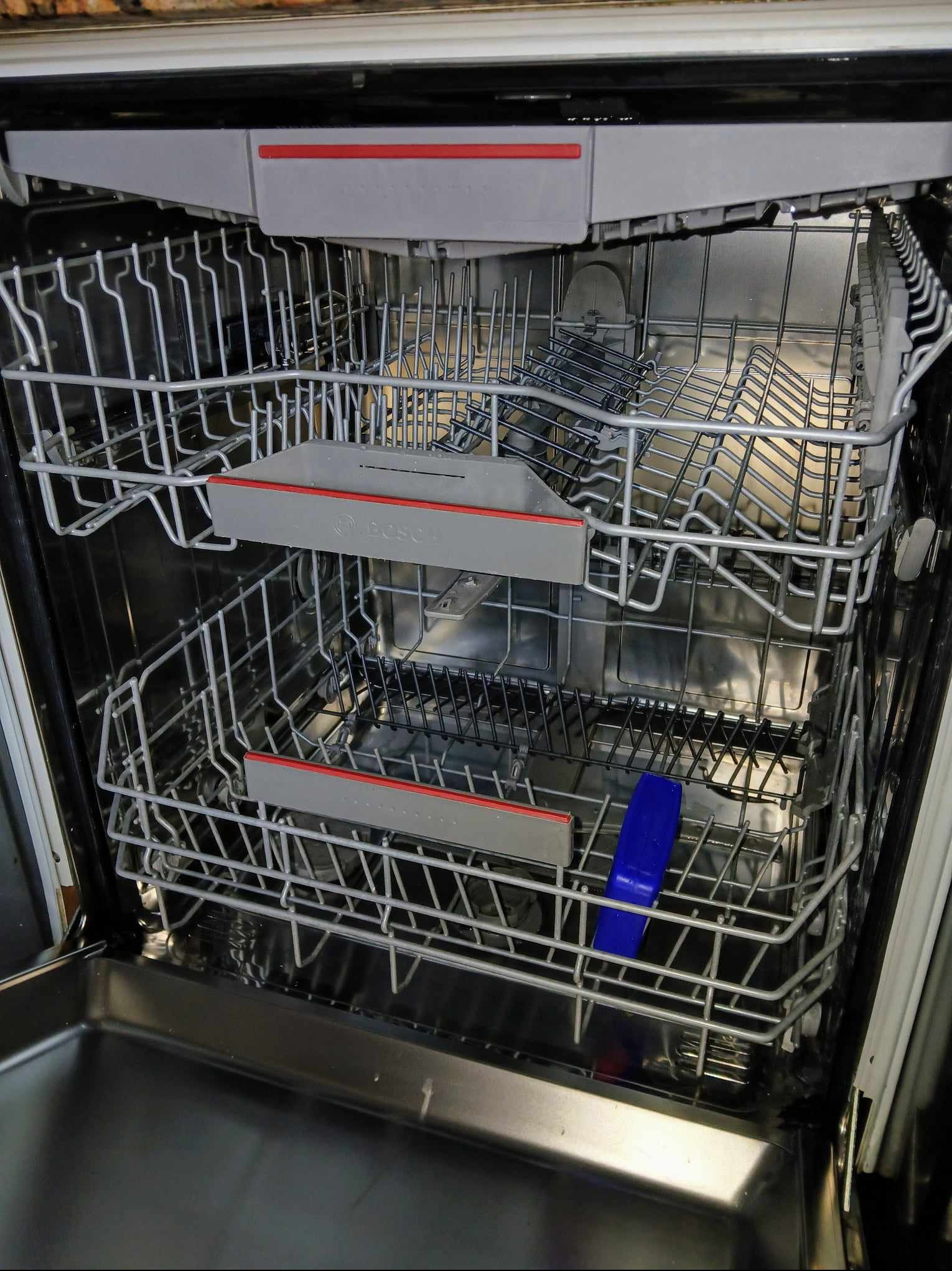 Wnętrze pustej zmywarki marki Bosch z dwoma wysuwanymi koszami na naczynia i niebieskim skrobakiem w dolnym koszu.