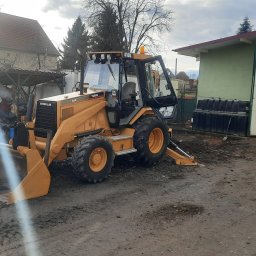 KRIS-POL LOGISTIC Anna Korecka - Żółta koparko-ładowarka Caterpillar 416B na błotnistym terenie, z otwartą kabiną i widoczną łyżką, zaparkowana przed budynkiem gospodarczym z otwartymi drzwiami, w tle dom i drzewa.