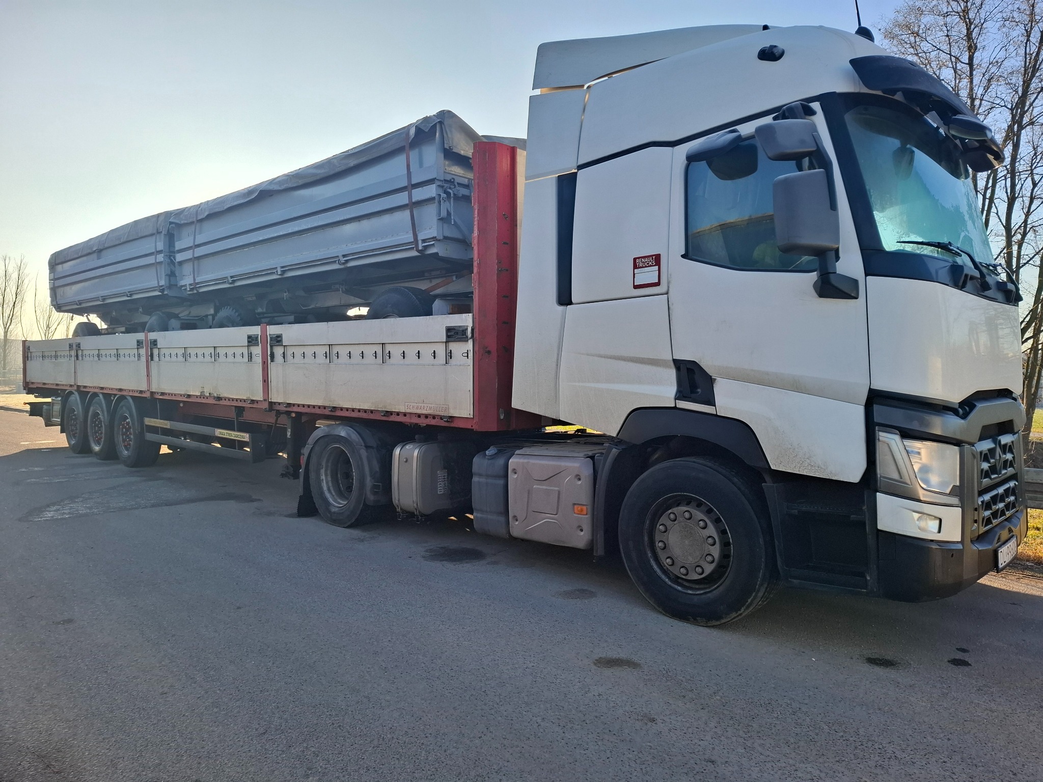 Biała ciężarówka Renault z naczepą przewożąca dwie szare, zakryte plandekami skrzynie ładunkowe, stojąca na asfaltowej drodze w słoneczny dzień.