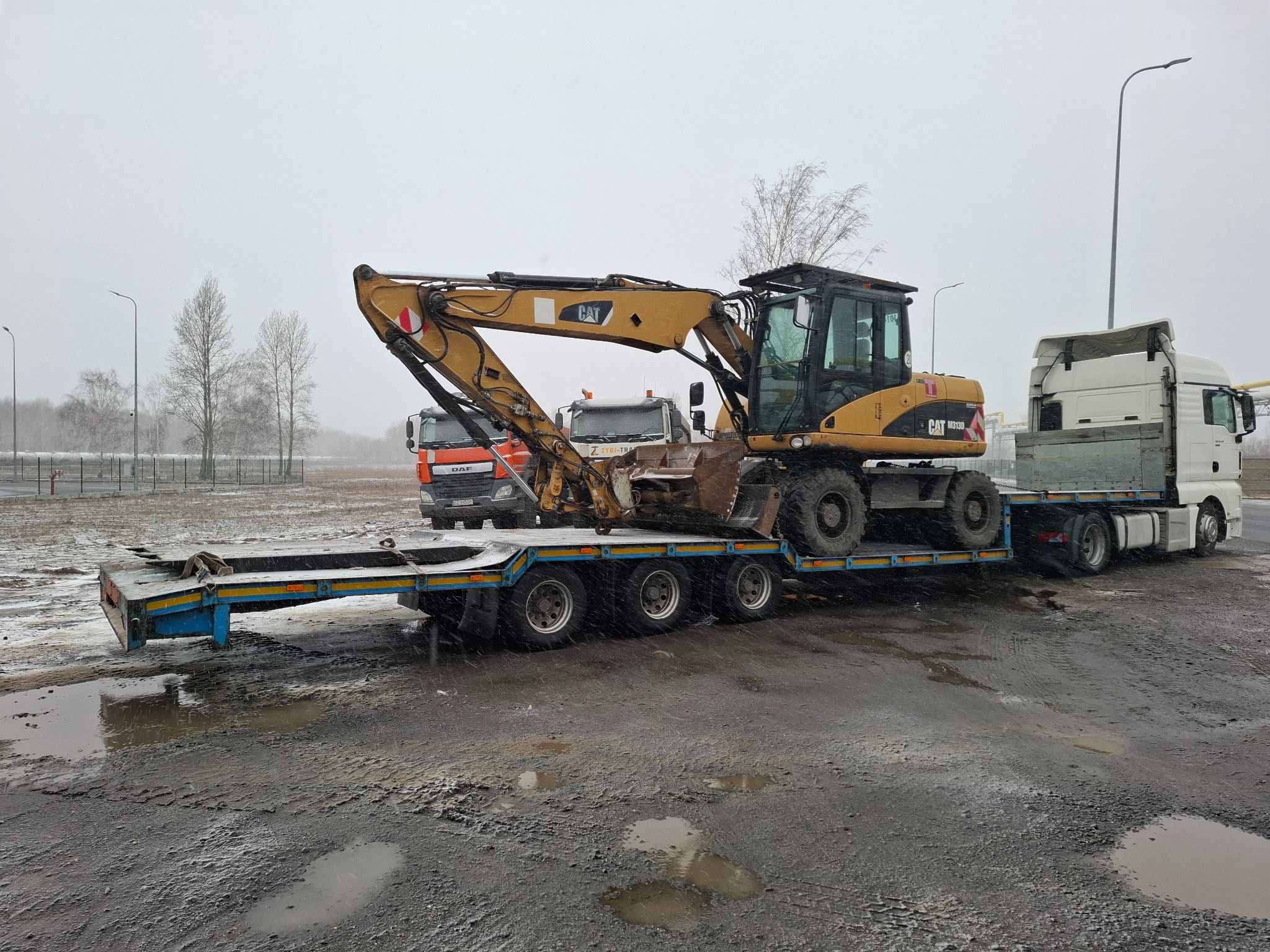 Żółta koparka CAT M313D na niebieskiej niskopodwoziowej przyczepie transportowana przez biały ciągnik siodłowy MAN, w tle widoczne inne pojazdy i ośnieżone drzewa.