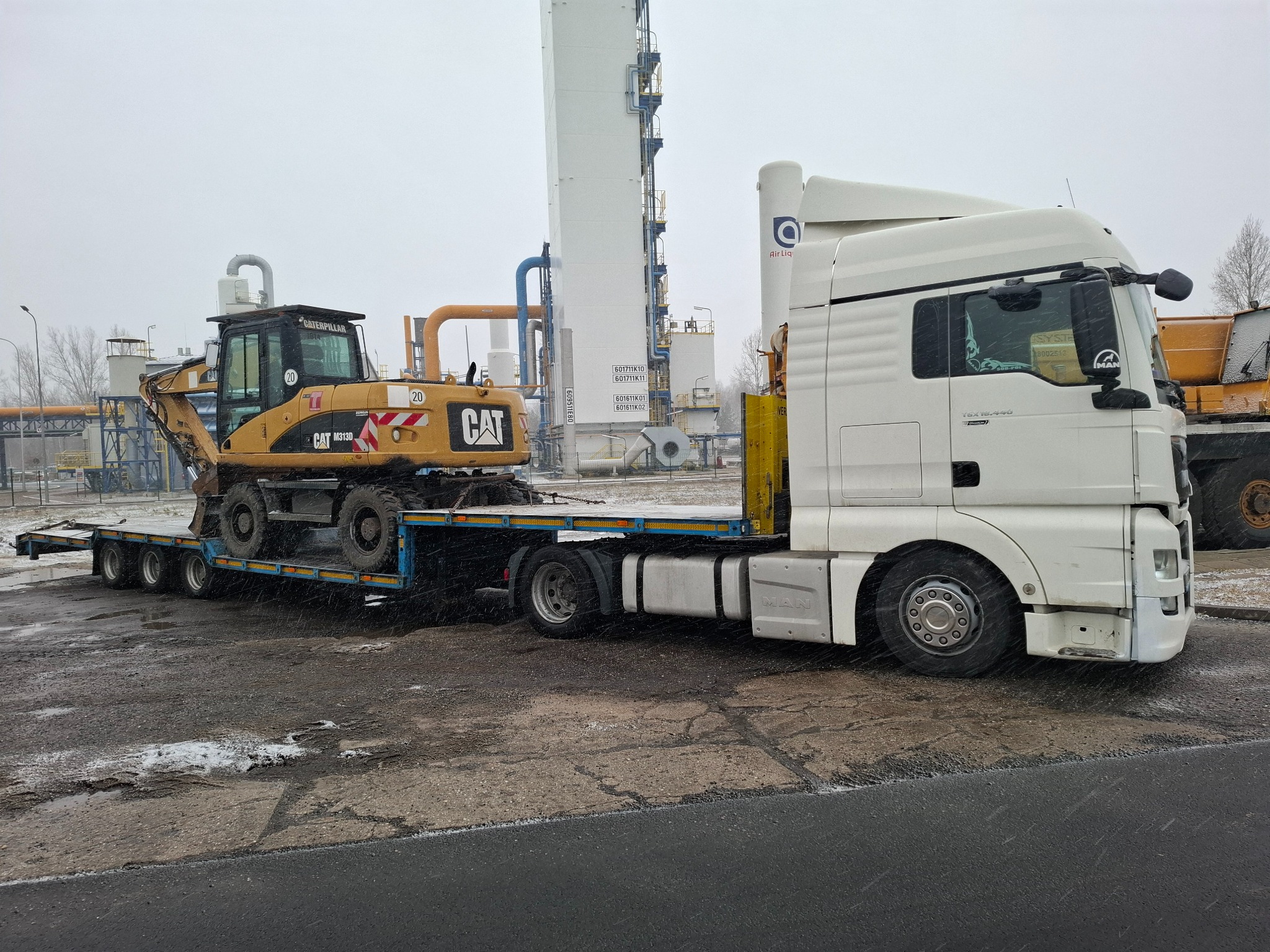 Biała ciężarówka MAN z naczepą przewożąca żółtą koparkę kołową CAT M313D na tle przemysłowego kompleksu z instalacjami.