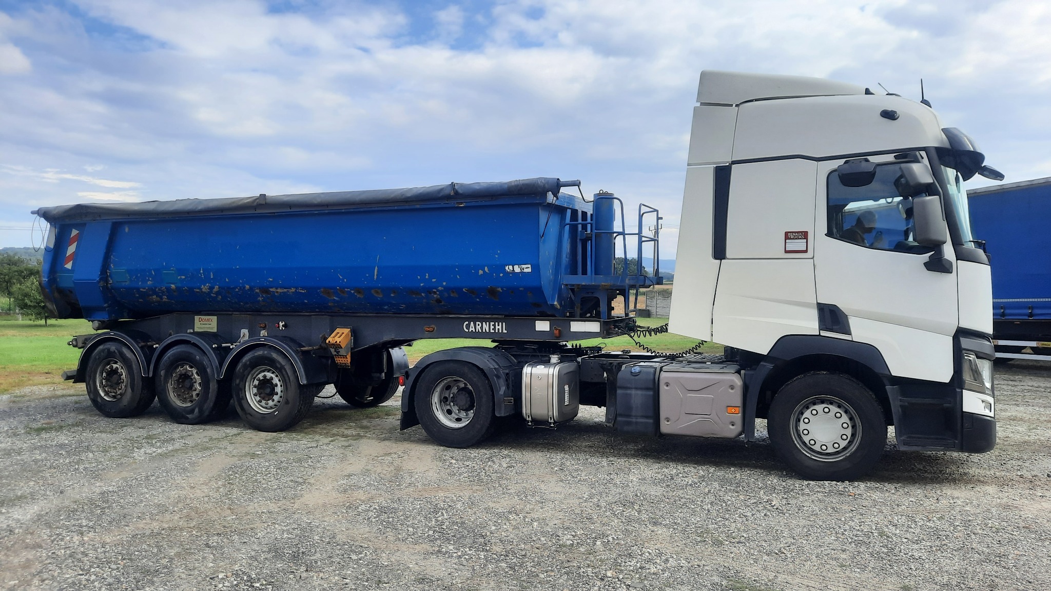 Ciężarówka z niebieską naczepą typu wywrotka marki Carnehl, stojąca na żwirowym terenie, pod błękitnym niebem z chmurami.