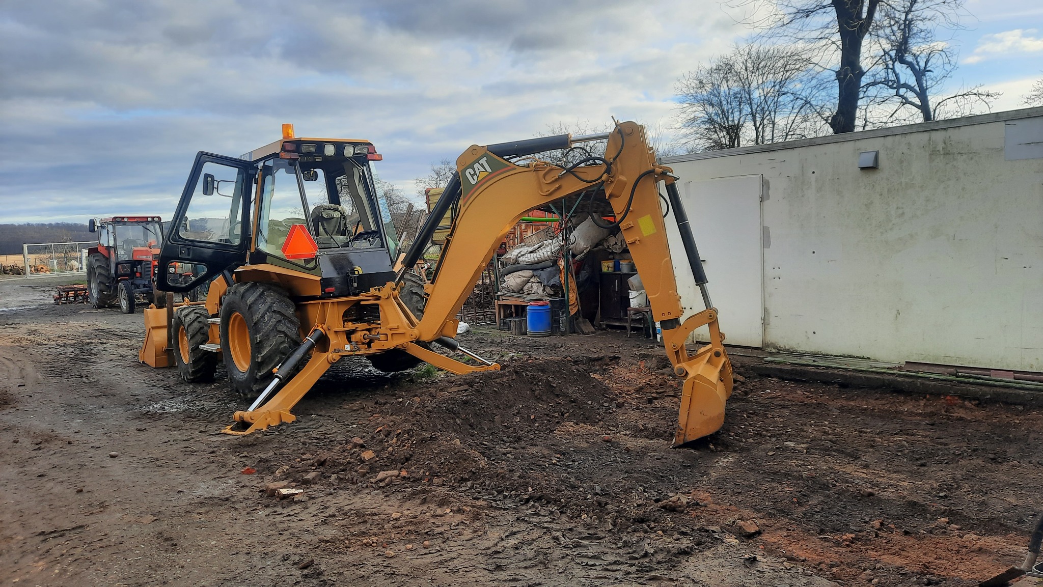 Żółto-czarna koparko-ładowarka CAT na nierównym, błotnistym terenie, z otwartą kabiną i widocznymi dwoma ciągnikami w tle, stojąca przed białą halą magazynową w pochmurny dzień.