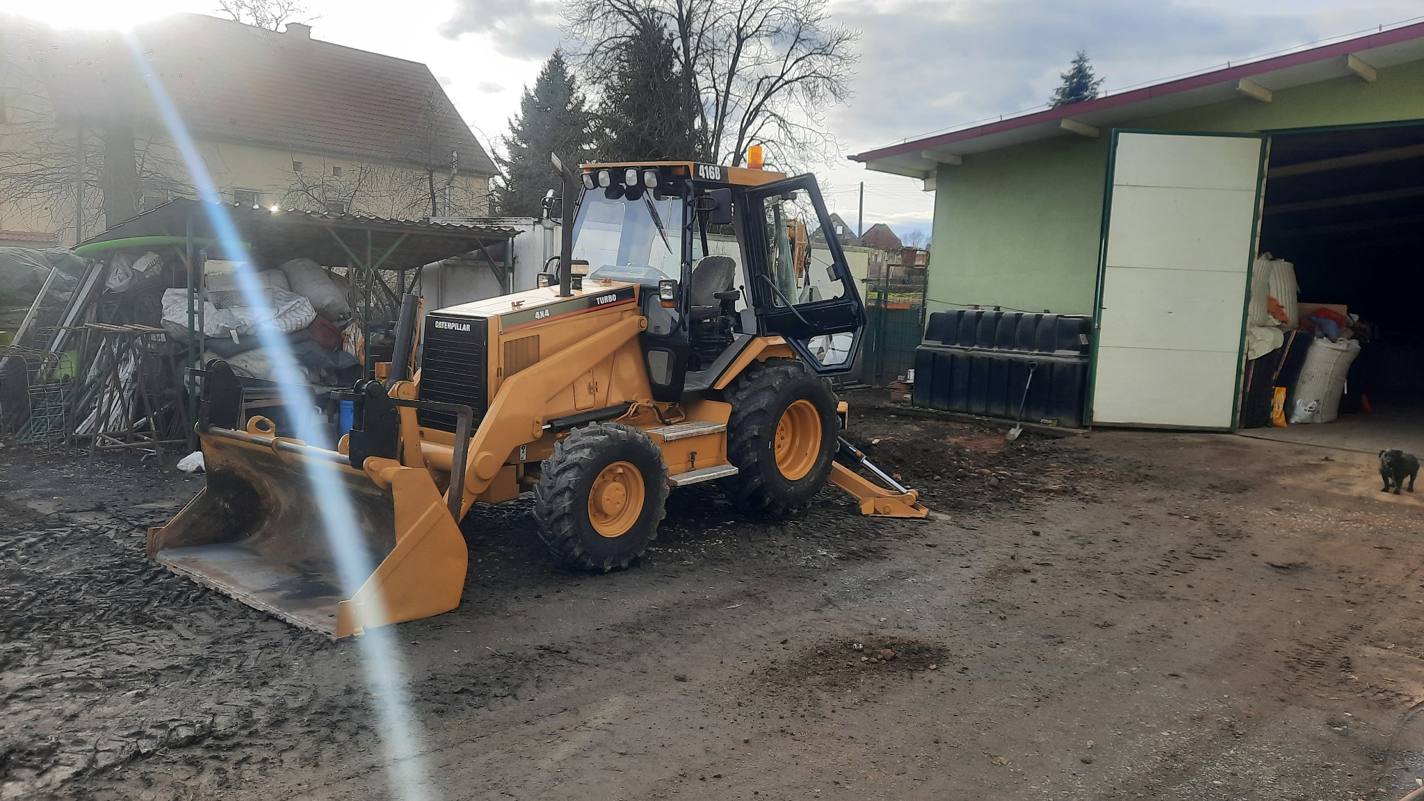 Żółta koparko-ładowarka Caterpillar 416B na błotnistym terenie, z otwartą kabiną i widoczną łyżką, zaparkowana przed budynkiem gospodarczym z otwartymi drzwiami, w tle dom i drzewa.