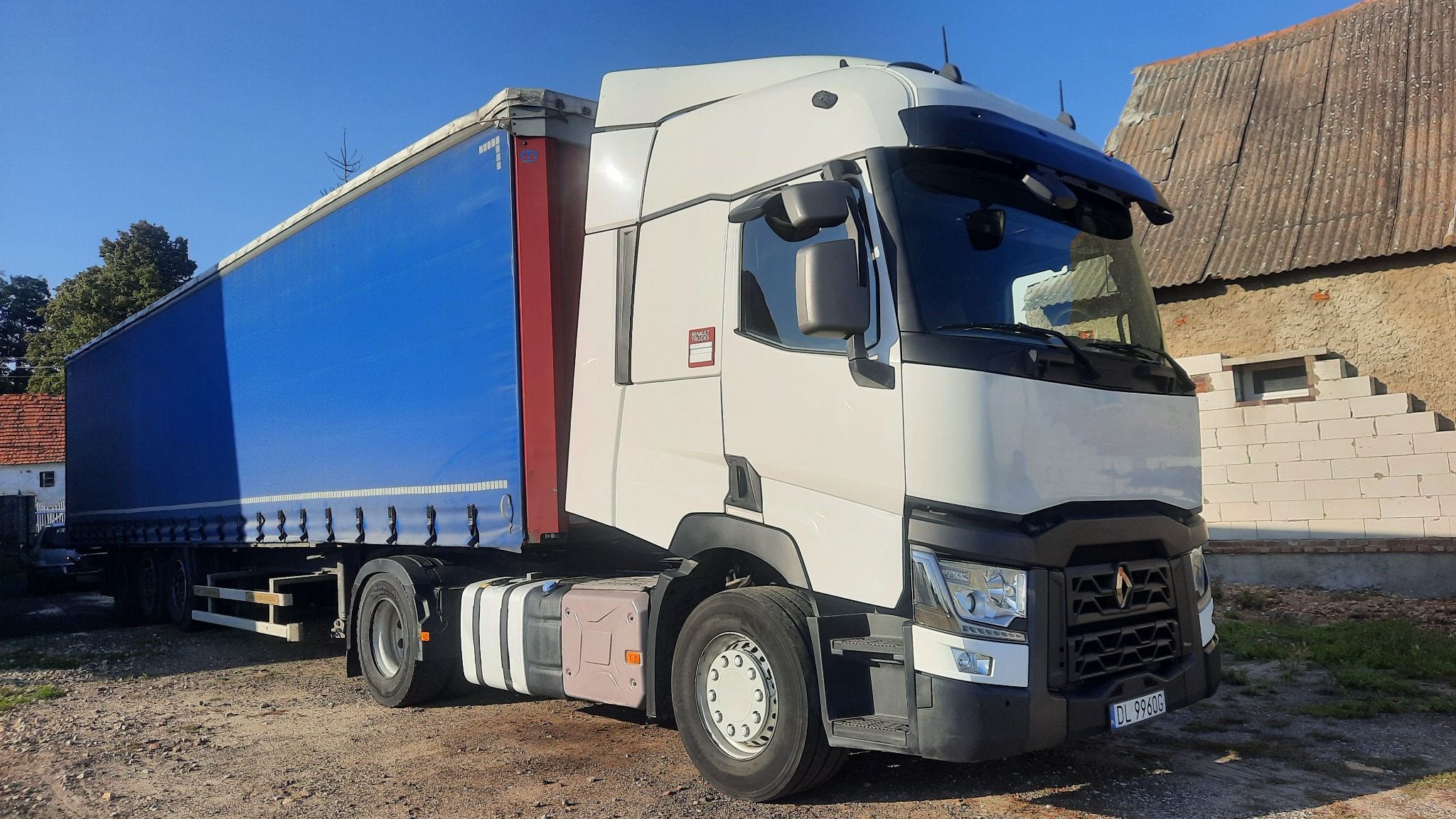 Ciężarówka Renault z niebieską plandeką zaparkowana na żwirowym podłożu, z widocznym budynkiem gospodarczym w tle i tablicą rejestracyjną DL 9960G.