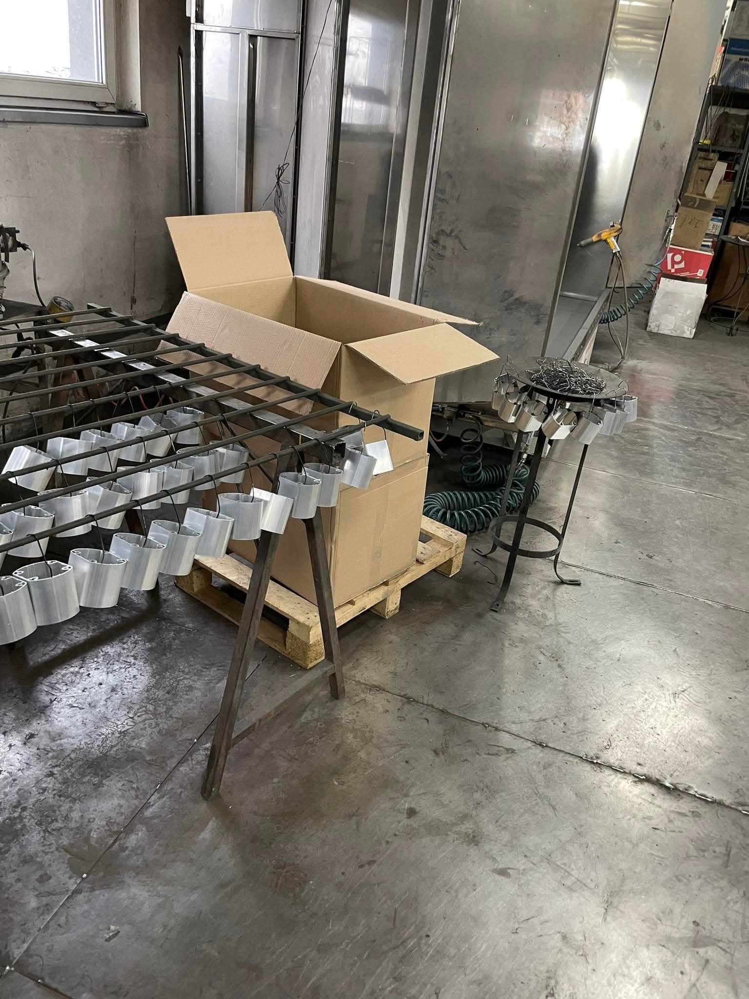 Metalowe elementy zawieszone na stojakach, przygotowane do procesu wykończeniowego. W tle widoczna komora lakiernicza i kartonowe pudła.