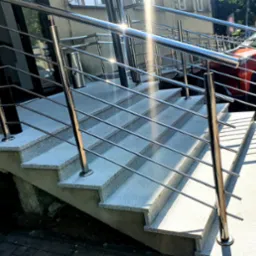 Schody wejściowe z szarego lastryko, zabezpieczone polerowaną konstrukcją z metalu, widoczne odbicia światła na powierzchni.