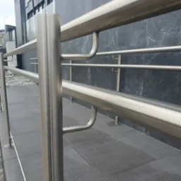 Dwu-poziomowa barierka ze stali nierdzewnej z prostymi słupkami i uchwytami, zamontowana na szarym chodniku przed budynkiem z ciemnoszarą elewacją.