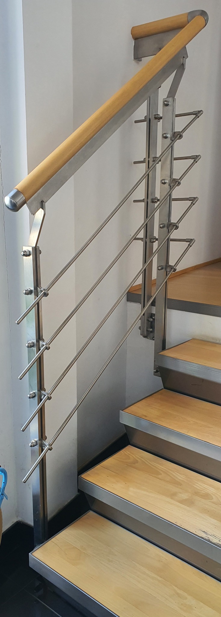 Nowoczesne schody z drewnianymi stopniami i balustradą z poziomych, cienkich prętów ze stali nierdzewnej, mocowanych do słupków za pomocą śrub.