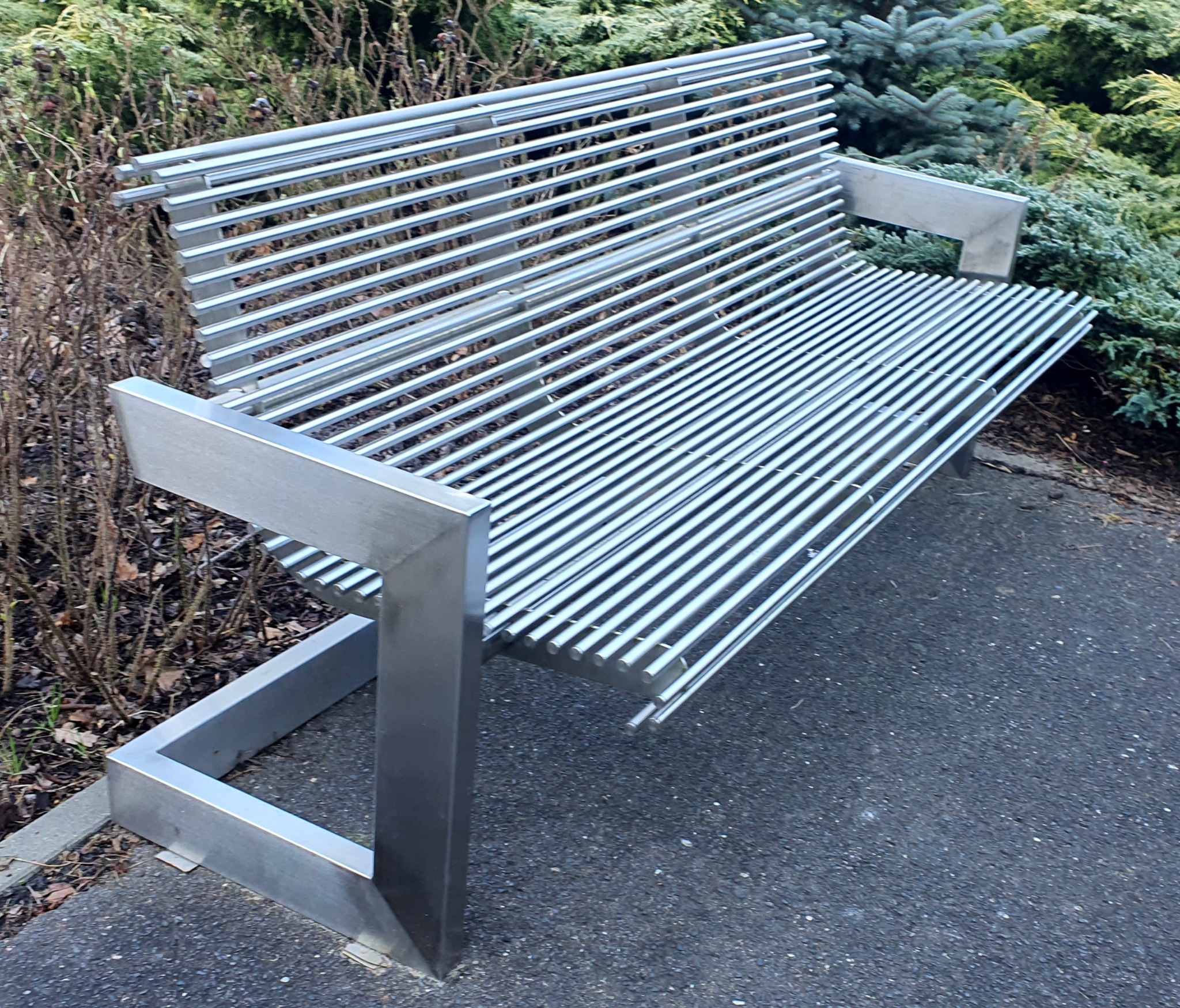 Nowoczesna ławka parkowa wykonana z polerowanych, metalowych prętów na tle zieleni.