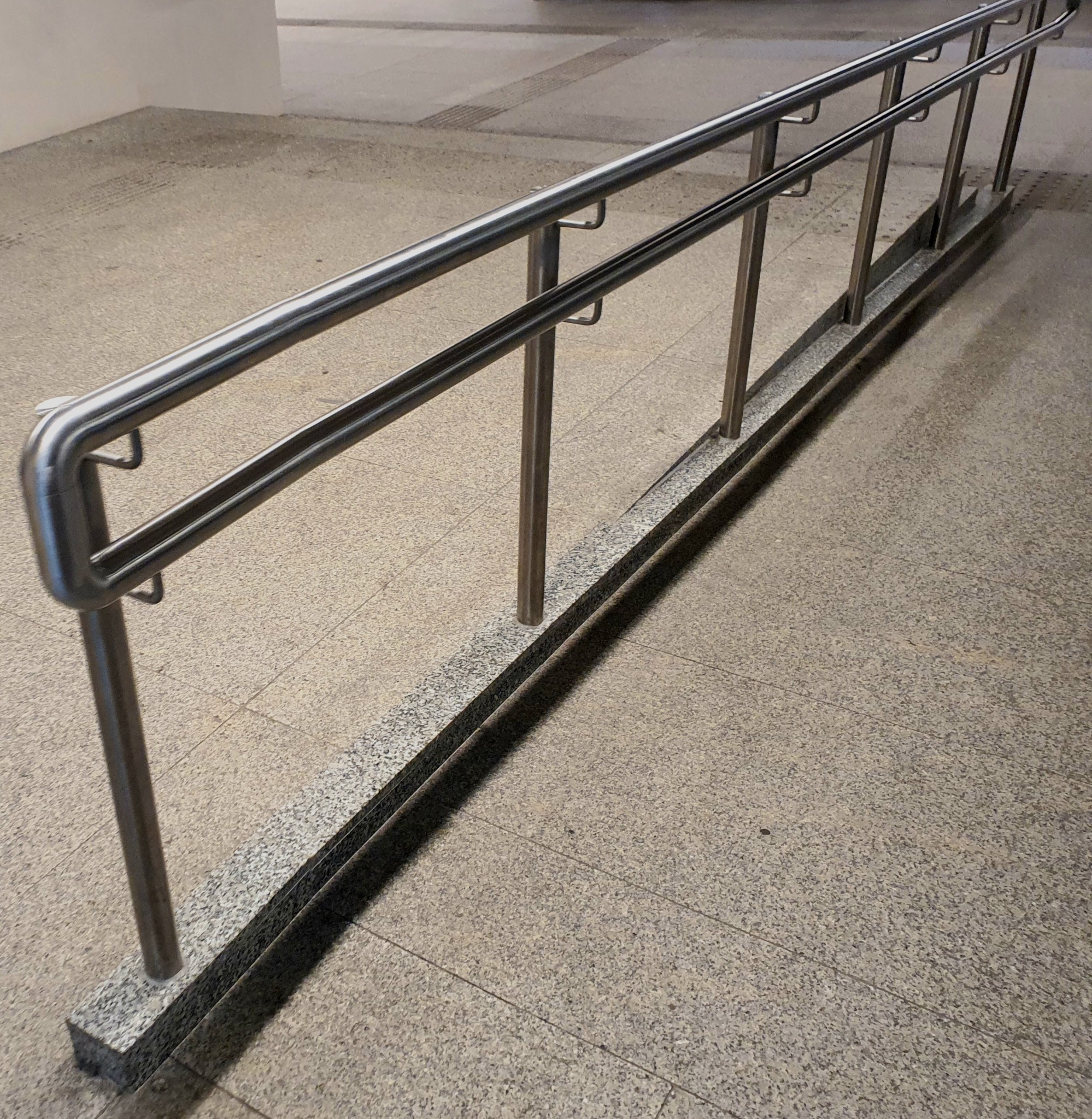 Szczegółowe ujęcie metalowej poręczy z uchwytami zamontowanej na niskim, granitowym murku, na tle kamiennej posadzki.