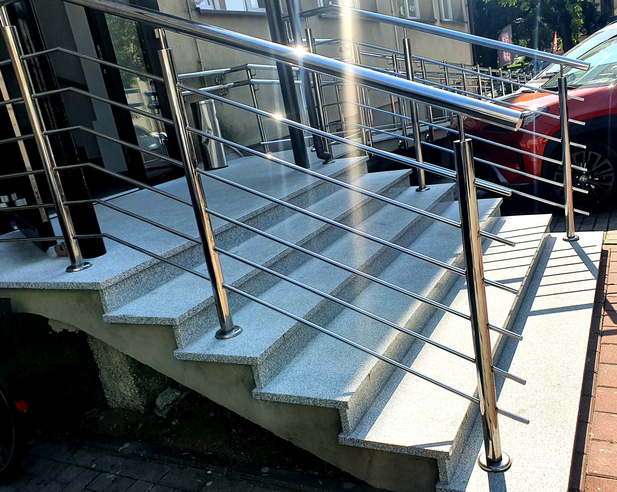 Schody wejściowe z szarego lastryko, zabezpieczone polerowaną konstrukcją z metalu, widoczne odbicia światła na powierzchni.