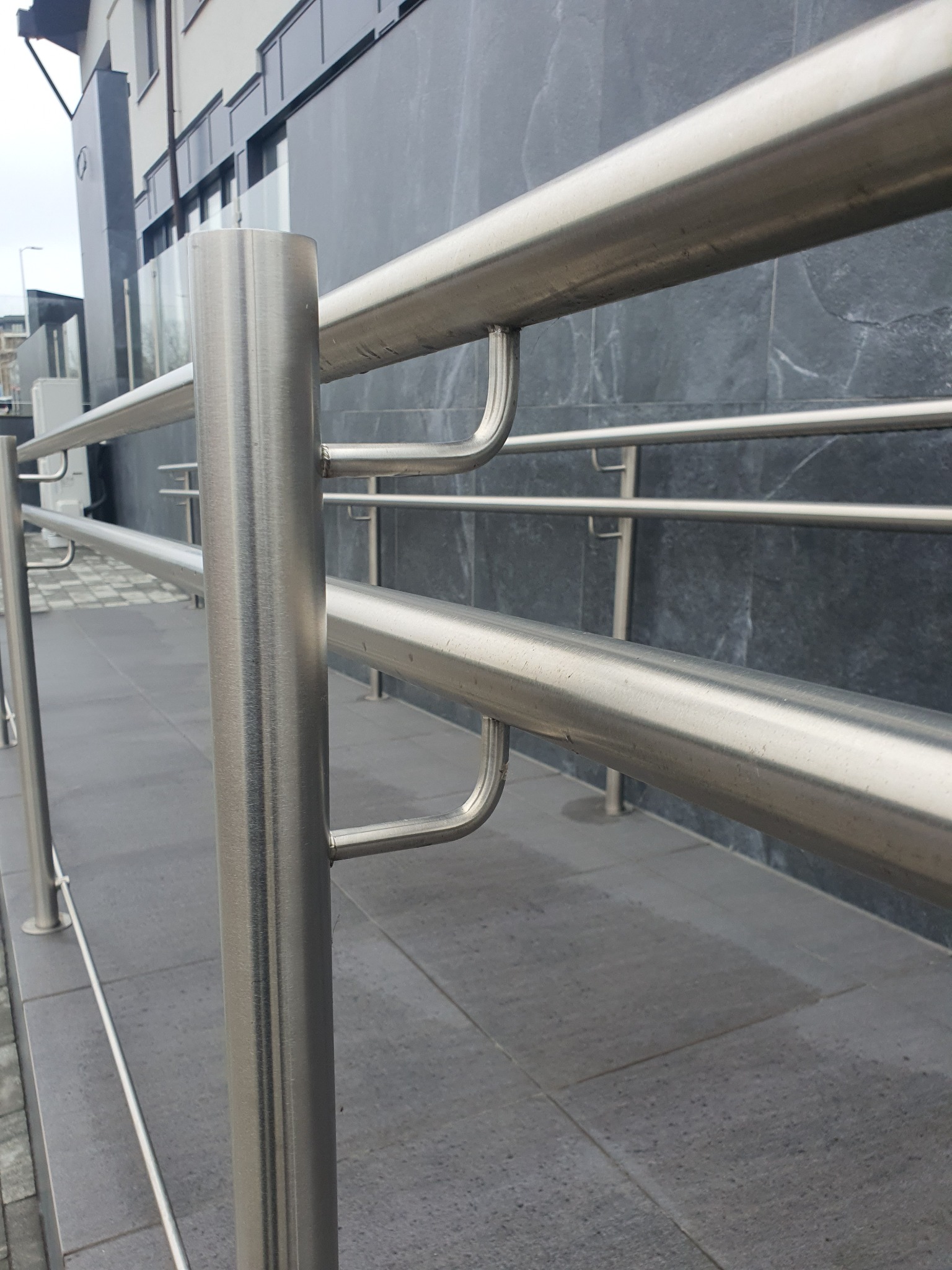 Dwu-poziomowa barierka ze stali nierdzewnej z prostymi słupkami i uchwytami, zamontowana na szarym chodniku przed budynkiem z ciemnoszarą elewacją.