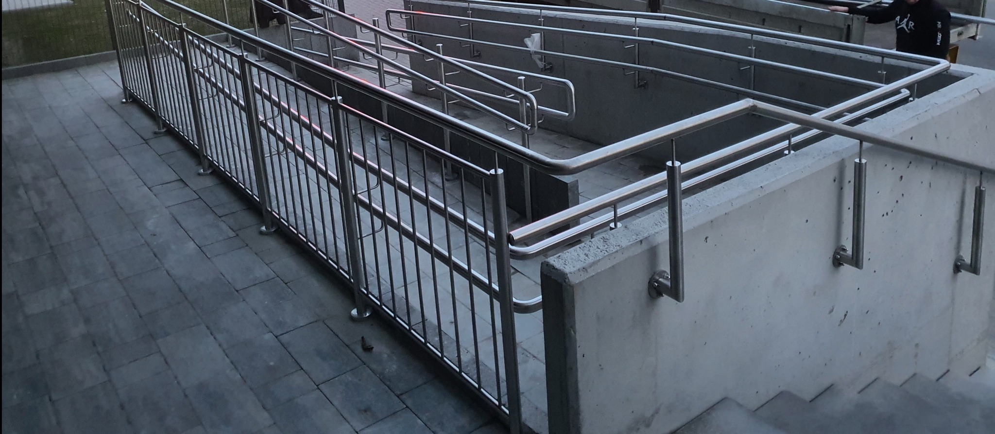 Zewnętrzne schody i rampa z betonowymi ścianami, zabezpieczone metalowymi, chromowanymi barierkami i poręczami, zapewniające dostęp do budynku.