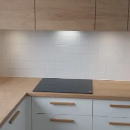 Nowoczesna kuchnia z drewnianymi szafkami górnymi i blatem, białe szafki dolne z drewnianymi uchwytami, płyta indukcyjna na blacie, białe kafelki na ścianie.