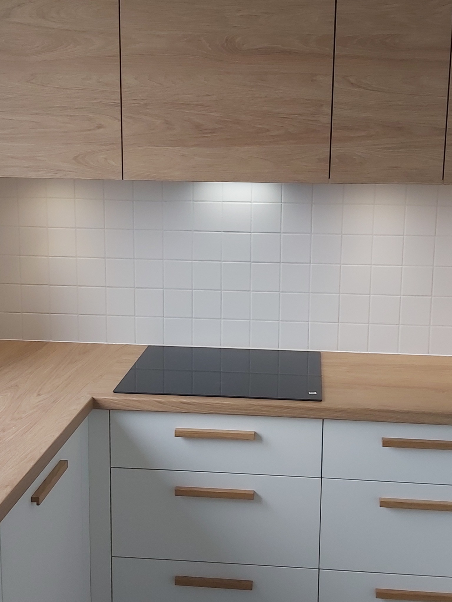 Nowoczesna kuchnia z drewnianymi szafkami górnymi i blatem, białe szafki dolne z drewnianymi uchwytami, płyta indukcyjna na blacie, białe kafelki na ścianie.