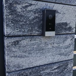 Bogdan Popłoński Clever-Tech - Czarny wideodomofon Ezviz zamontowany na szarym, kamiennym słupku ogrodzeniowym. Widoczny obiektyw kamery i przycisk dzwonka. Słoneczny dzień.