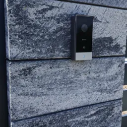 Czarny wideodomofon Ezviz zamontowany na szarym, kamiennym słupku ogrodzeniowym. Widoczny obiektyw kamery i przycisk dzwonka. Słoneczny dzień.