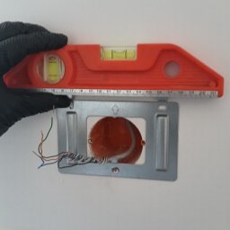 Bogdan Popłoński Clever-Tech - Instalacja puszki elektrycznej w ścianie, poziomowana pomarańczową poziomicą. Widoczne przewody i ręka w czarnej rękawicy.